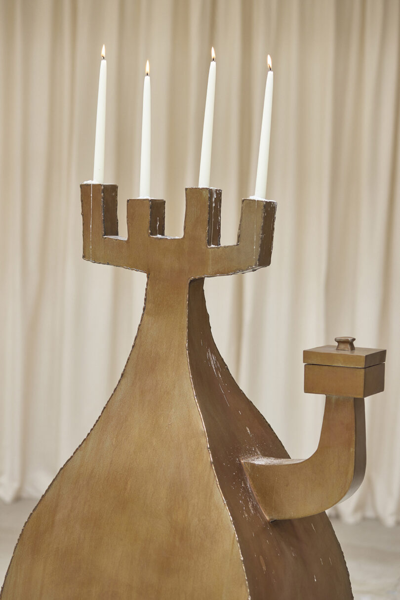 A sculptural candelabrum with four lit white candles and a box-like extension, set against a neutral, draped background.