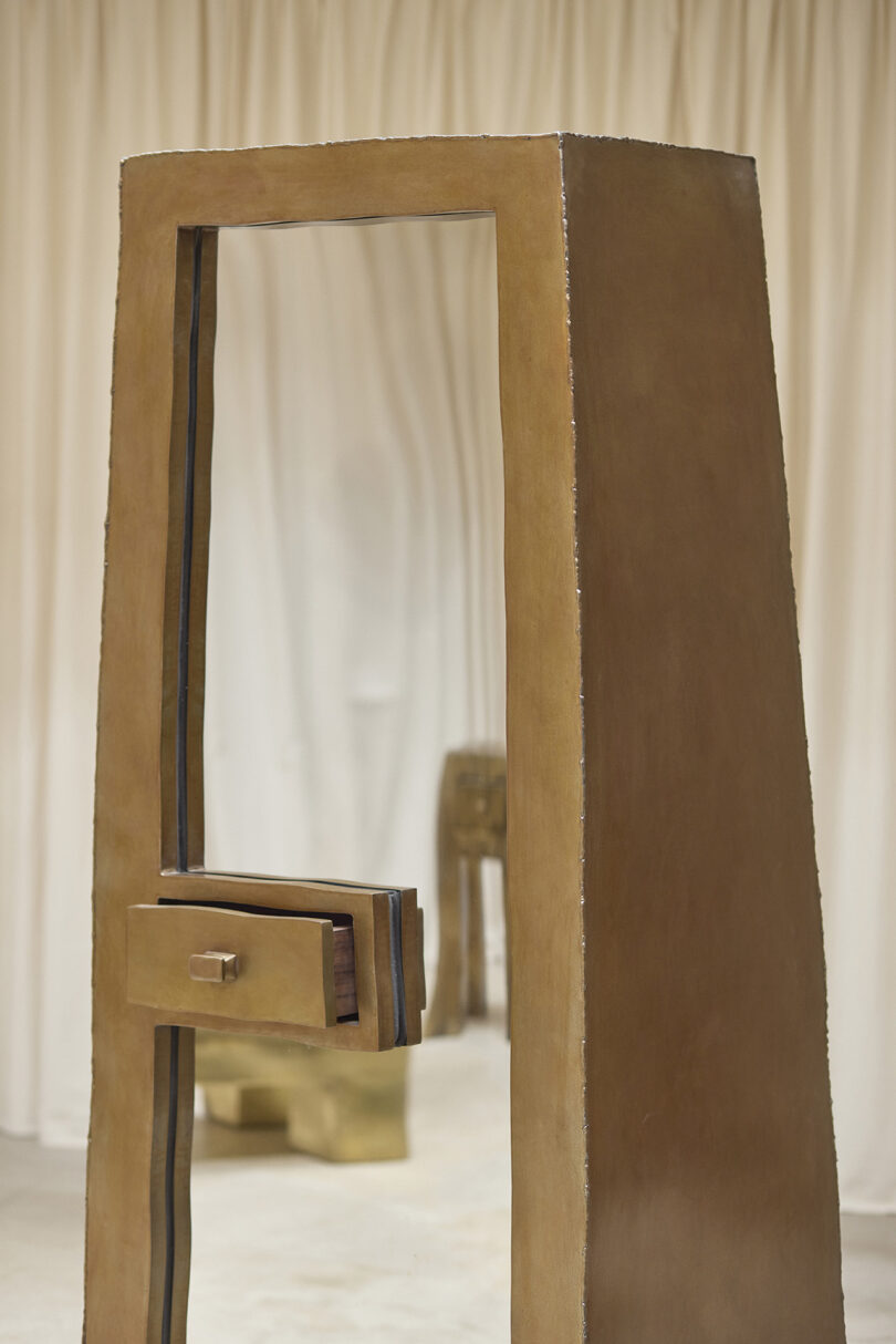 A geometric, brown sculptural structure stands upright with an open rectangular frame and protruding panel, set against a neutral, draped background.
