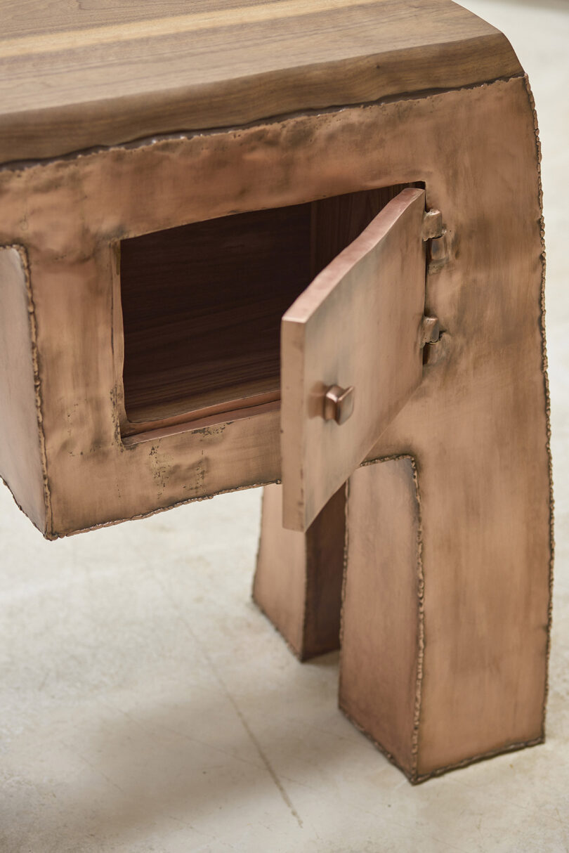 A close-up of a metallic side table with a rectangular cabinet door partially open, revealing a wooden interior. The table has a copper finish and visible welded seams.