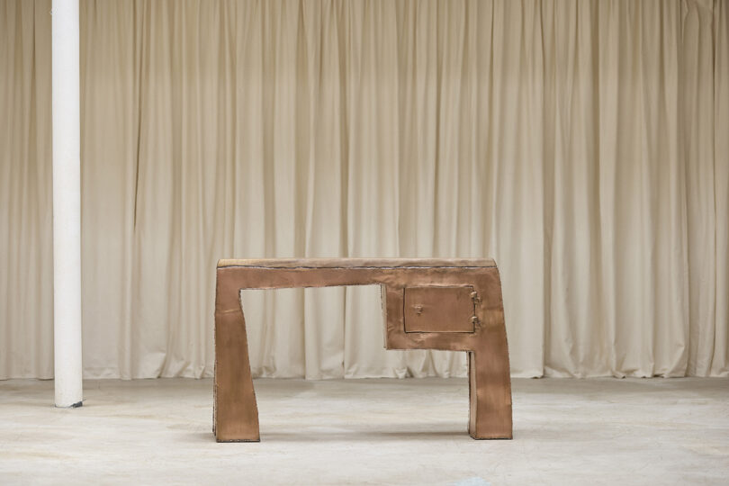 A metallic, geometric table with an asymmetric design sits on a concrete floor in front of beige curtains.