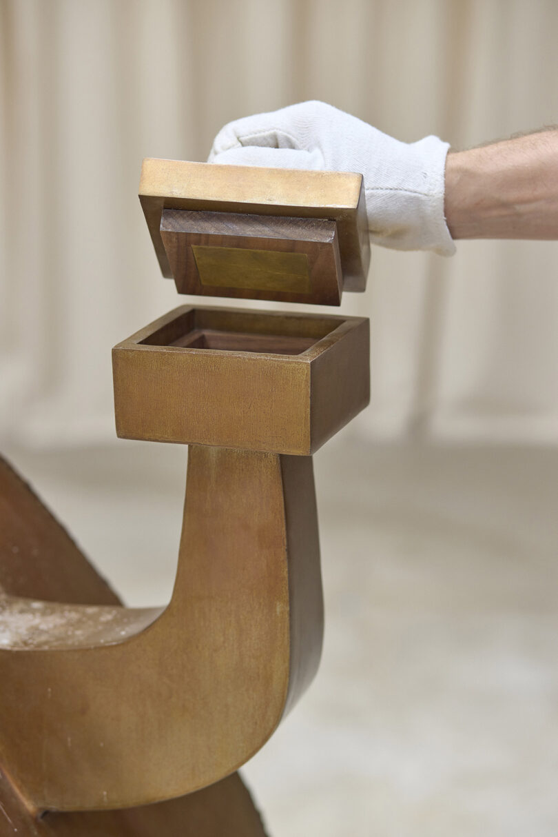 A gloved hand opens a small, square, wooden box attached to a curved wooden stand against a neutral background.
