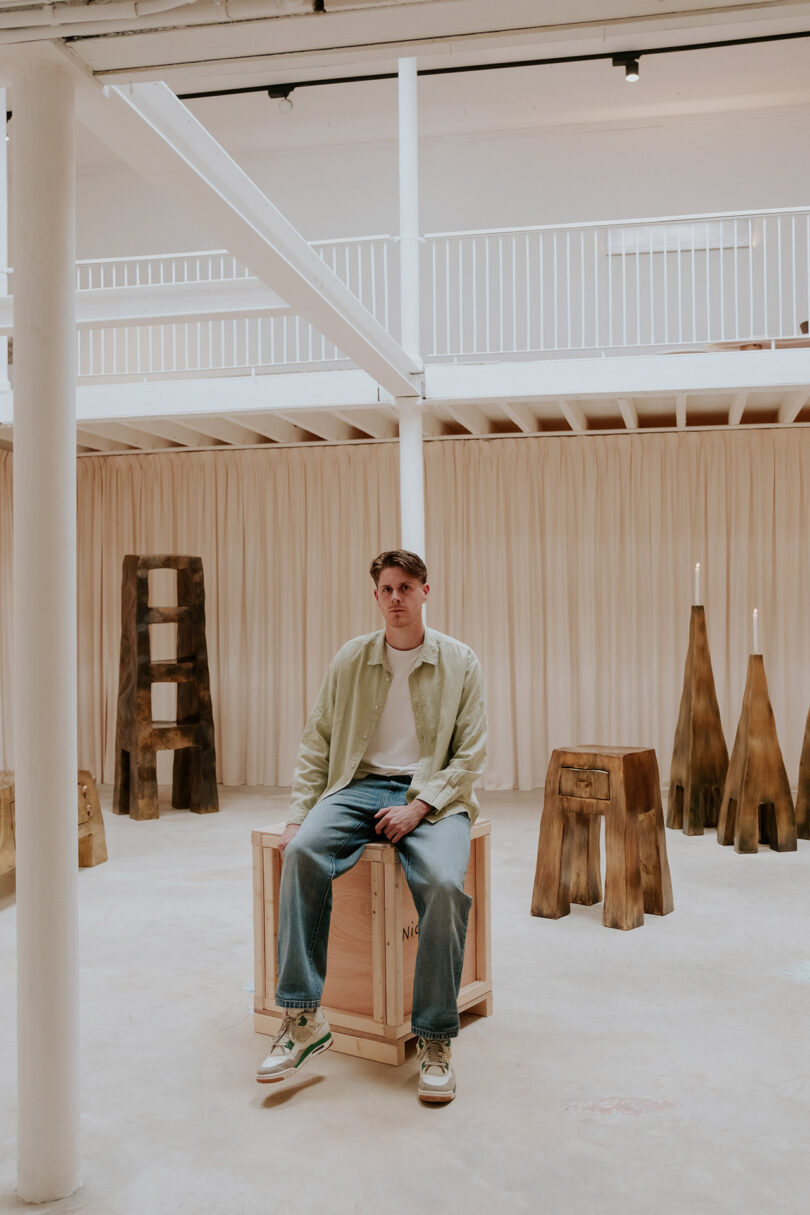 A man in casual clothing sits on a wooden crate in a spacious room with wooden sculptures and a curtained backdrop.