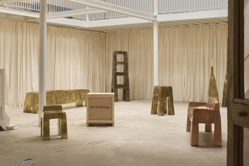 A spacious room with light beige curtains and floor, displaying various metallic and wooden sculptural furniture pieces and a crate labeled "Nick Winton.
