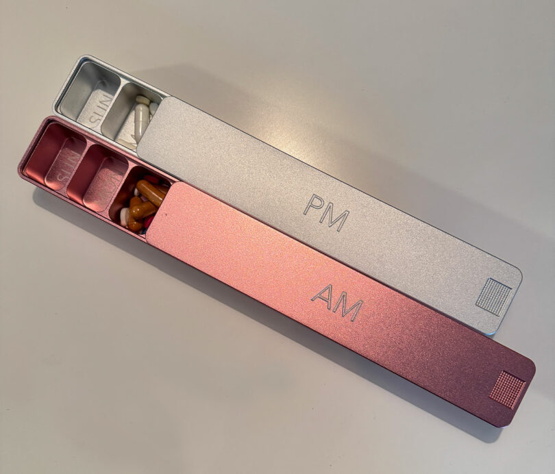 Two pill organizers labeled "AM" and "PM" contain various pills and capsules, placed on a white surface. The "AM" section holds more pills than the "PM" section.