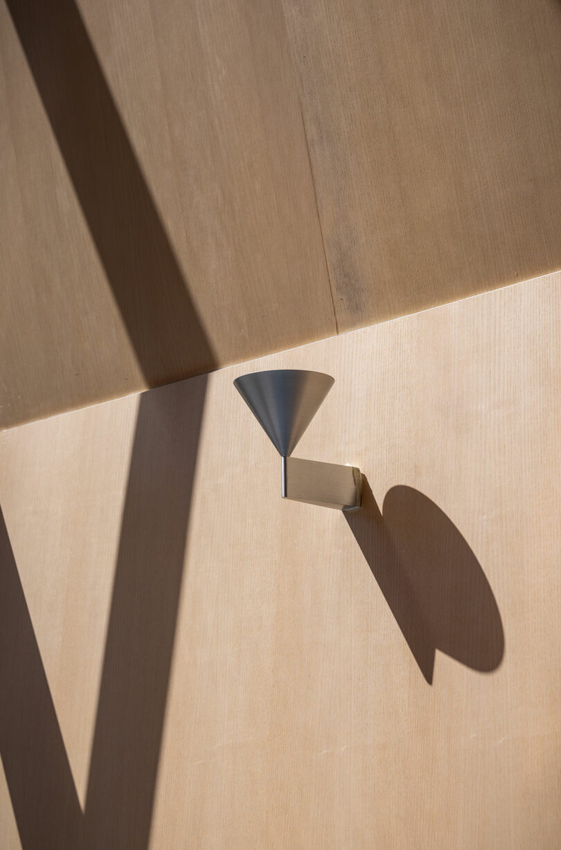 A modern, cone-shaped wall sconce casts a distinct shadow on a wooden wall illuminated by angled sunlight.