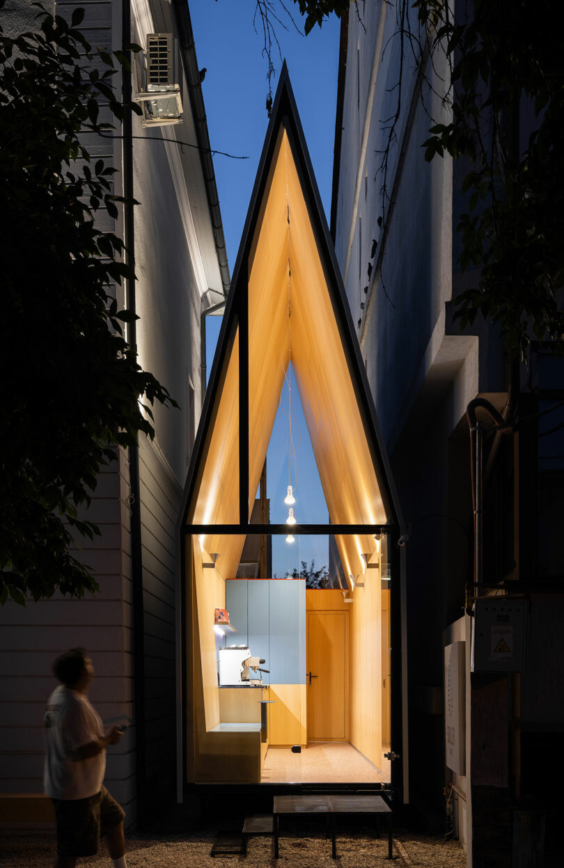 A-frame micro house with a glass front, wood interior, and modern lighting, nestled between two buildings at dusk. A person walks by in the foreground.