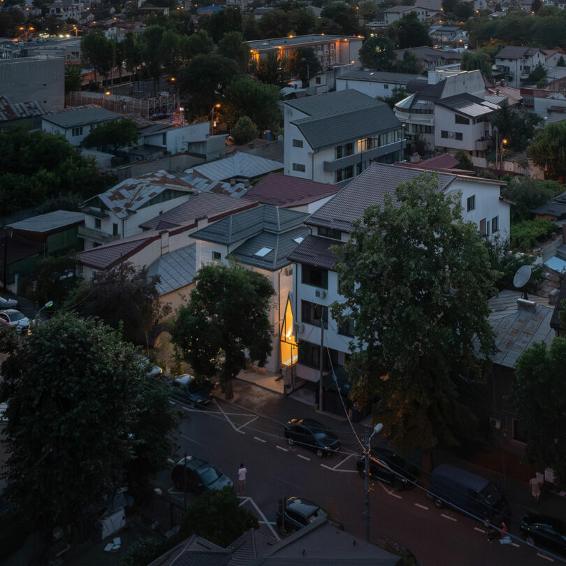 Aerial view of a residential neighborhood at dusk, featuring multi-story houses, parked cars along the street, and trees surrounding the buildings.