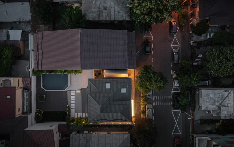 Aerial view of a residential neighborhood at dusk, showing houses, a driveway, streetlights, parked cars, and trees lining the street.