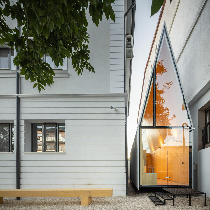 A modern narrow house with a sharp triangular roof and large orange-tinted glass facade is built between two older white buildings. A wooden bench is in the foreground.