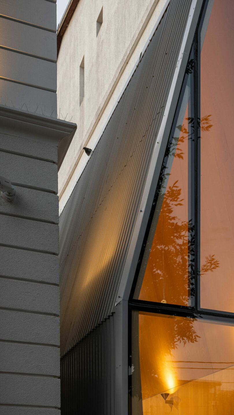 Close-up of two adjacent building facades, one with white horizontal siding and the other with dark metal panels and large orange-tinted glass windows reflecting light and tree branches.
