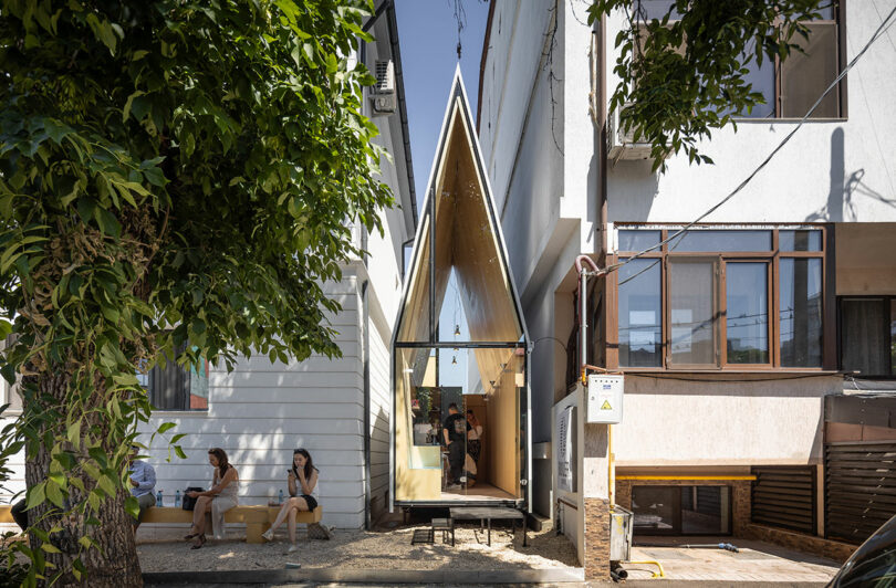 A narrow, modern building with a sharply pointed roof stands between two larger buildings; people are seated outside under a tree.