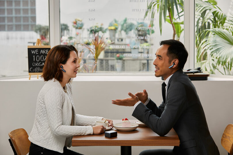 Two people sit at a café table, both wearing wireless earbuds, having a conversation; a coffee cup and dessert plate are on the table.