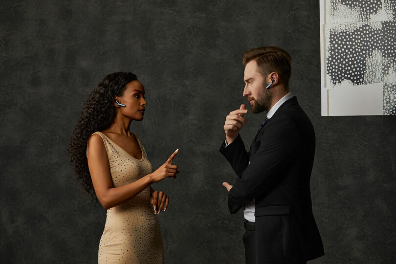 A woman in a beige dress and a man in a suit, both wearing wireless earpieces, stand facing each other and conversing against a dark textured background.