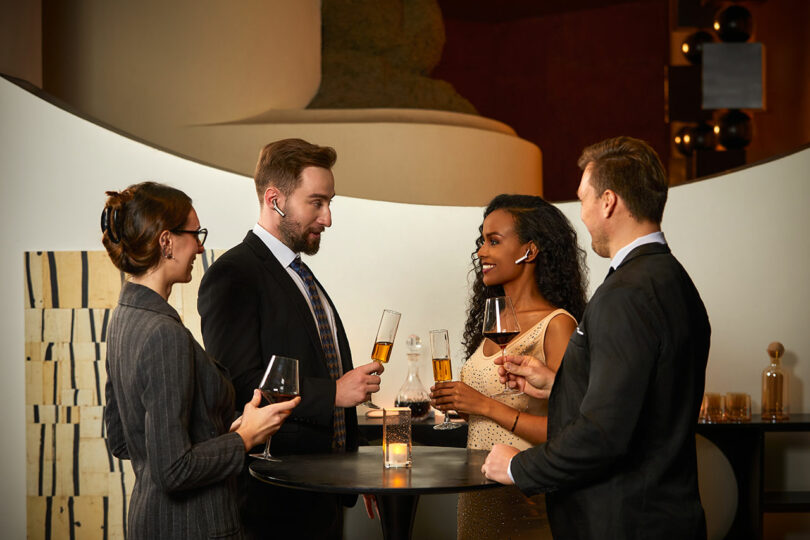 Four people dressed in formal attire stand around a tall table, holding drinks and conversing in a stylish indoor setting.