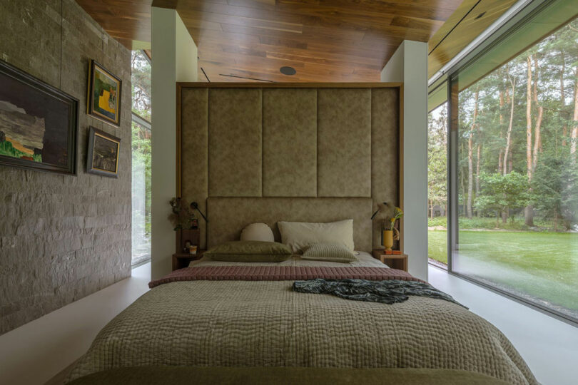 A modern bedroom with a large bed, textured headboard, stone accent wall, wood ceiling, floor-to-ceiling windows, and a garden view.