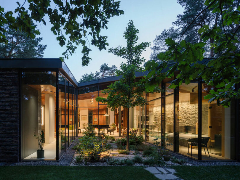 Modern house with large floor-to-ceiling glass windows, surrounding an illuminated courtyard garden, set among trees in the evening.