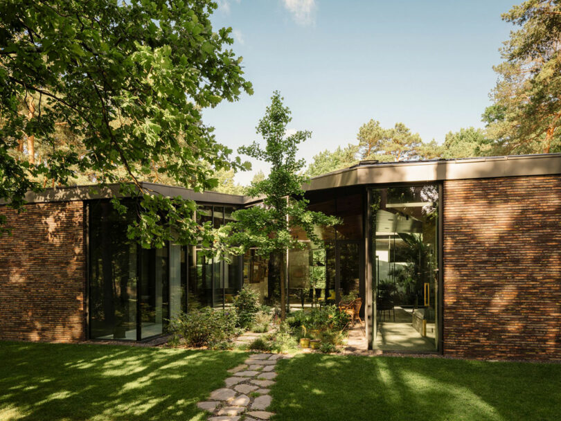 Modern house with large floor-to-ceiling windows, dark brick exterior, and a landscaped yard with stepping stones, surrounded by trees and greenery.
