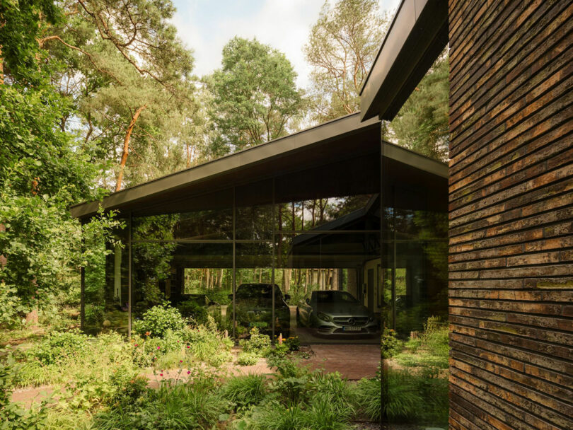 A modern house with floor-to-ceiling glass walls, a visible car parked inside, and surrounded by dense greenery and trees.
