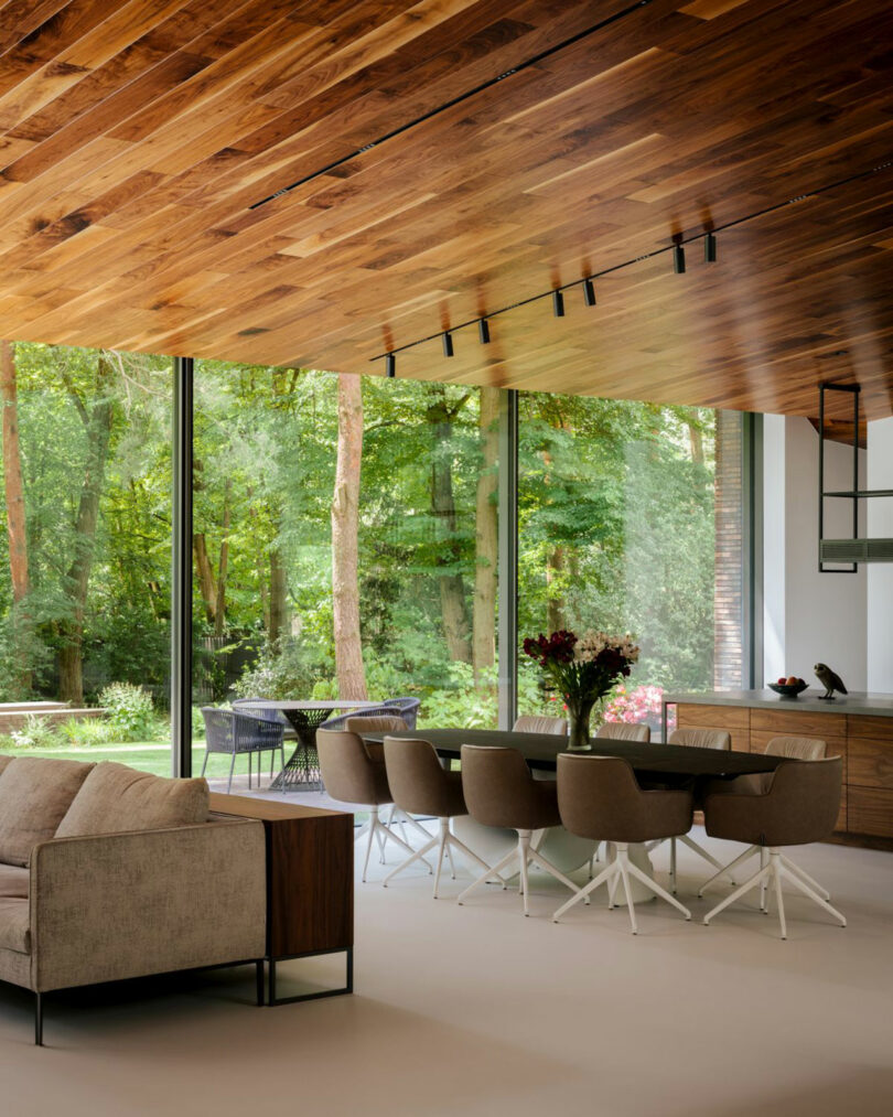 Modern living and dining area with floor-to-ceiling windows, wood ceiling, neutral furniture, and a view of a green forest outside.