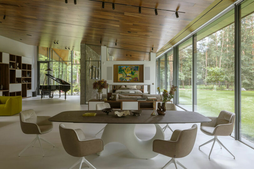 Modern open-concept living space with large windows, a dining table with chairs, a bed, bookshelves, a grand piano, and views of a green lawn and forest outside.