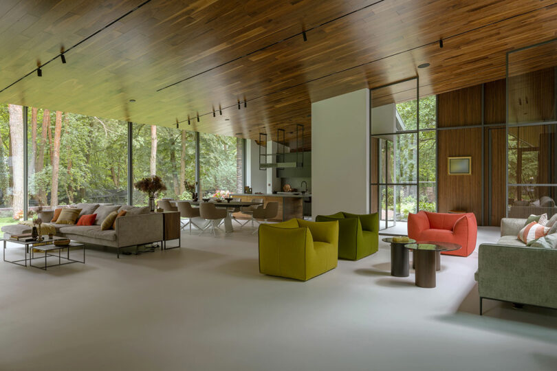 Spacious modern living room with large windows, wood ceiling, neutral sofas, green and orange armchairs, and views of a forest outside.
