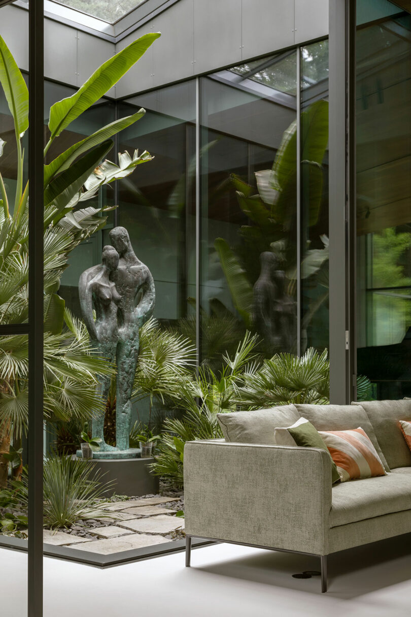 A modern glass-walled room with a green sofa faces an outdoor garden featuring a large abstract bronze sculpture surrounded by tropical plants.