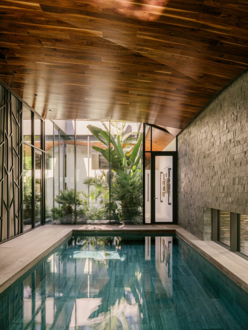 Indoor swimming pool with tiled floor, stone wall, wood ceiling, and large glass windows overlooking lush tropical plants in a courtyard.
