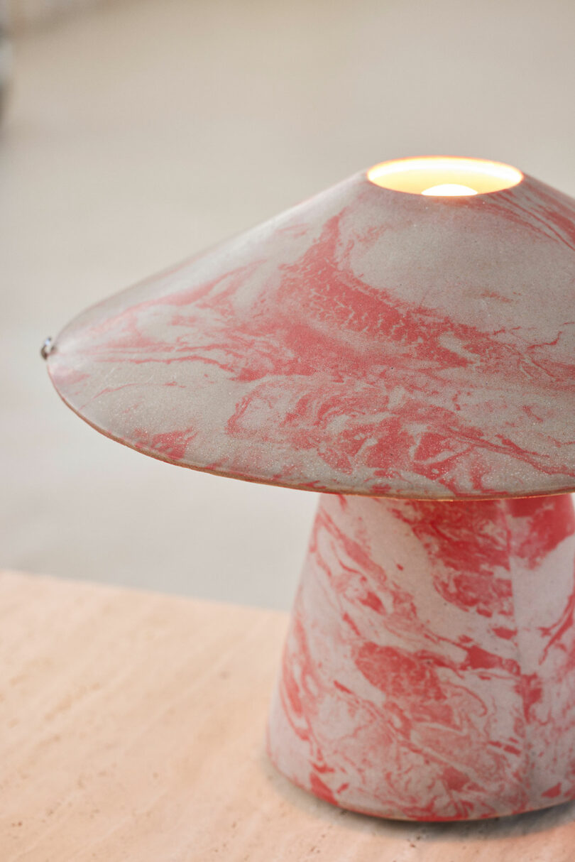 A pink and gray marbled Upglas Luca Nichetto table lamp with a wide, conical shade emits a warm light, placed on a light wood surface.