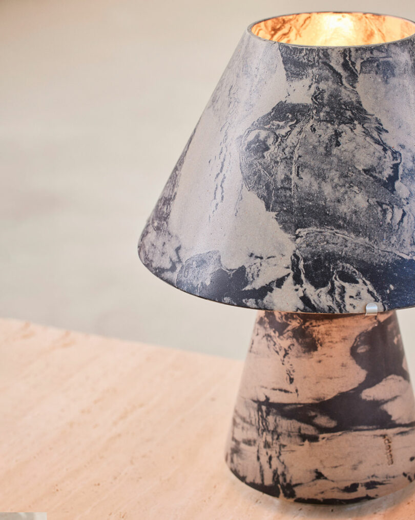 A stone-textured Upglas Luca Nichetto table lamp with a conical shade sits on a light-colored wooden surface.