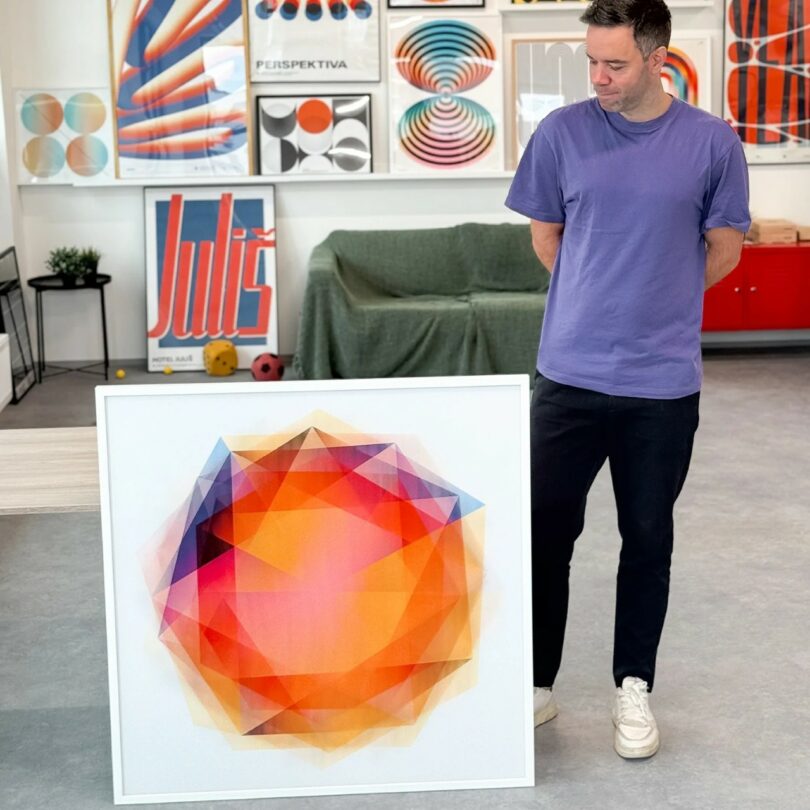 A man in a purple shirt stands indoors, looking at a large framed geometric artwork with warm orange, red, and yellow tones. Graphic posters are displayed on the wall behind him