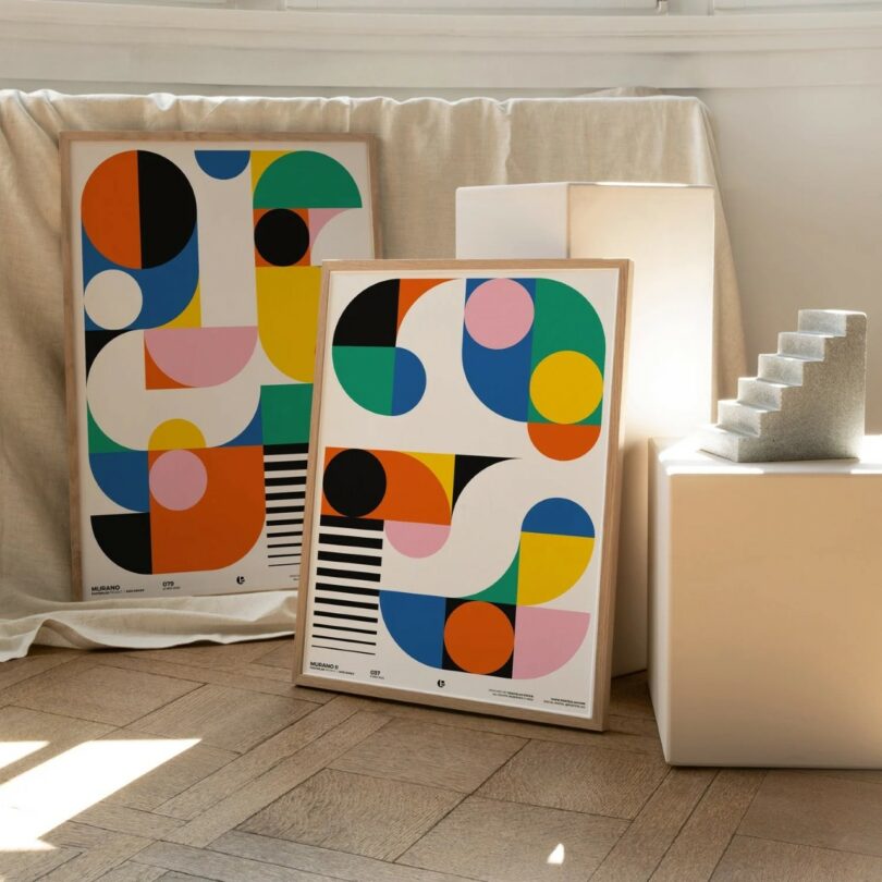 Two framed abstract art prints with colorful geometric shapes are displayed on the floor next to a small gray staircase sculpture and white blocks in a sunlit room