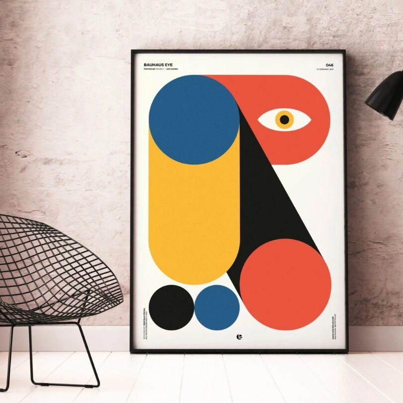 A framed abstract artwork with geometric shapes in red, blue, yellow, and black, featuring an eye motif, leaning against a light-colored wall next to a wireframe chair