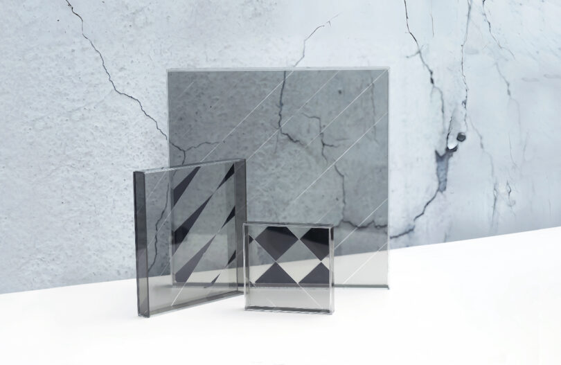 Three glass panels with diagonal and diamond-shaped patterns are displayed upright on a white surface against a cracked concrete wall background