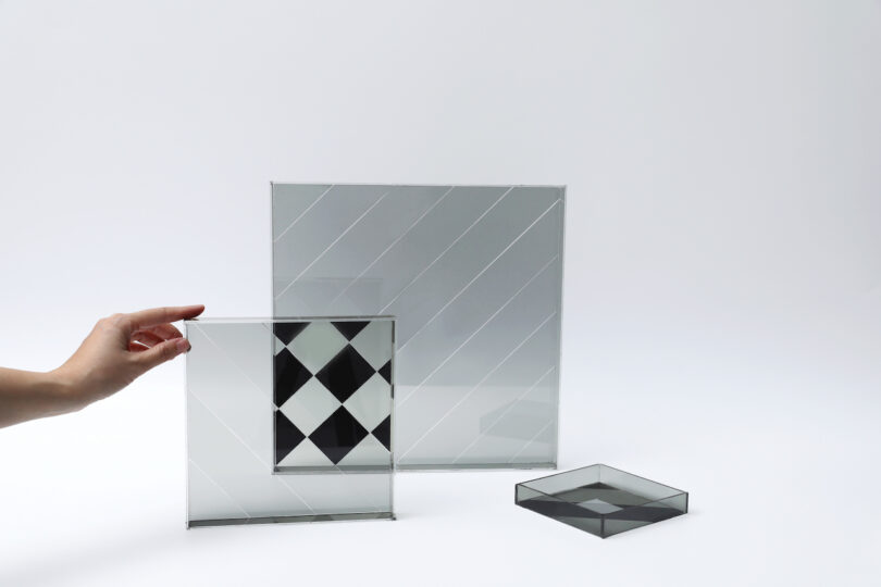 A hand holds a square glass pane with a black and white diamond pattern, next to two larger clear glass panes with diagonal lines, all against a plain white background