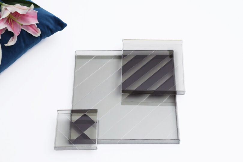 Three transparent, square, geometric trays with diagonal lines are arranged on a white surface near a blue cushion and pink lilies
