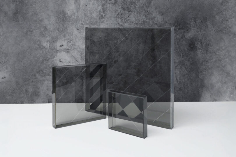 Three rectangular tinted glass panels of different sizes are standing upright on a white surface against a gray textured background