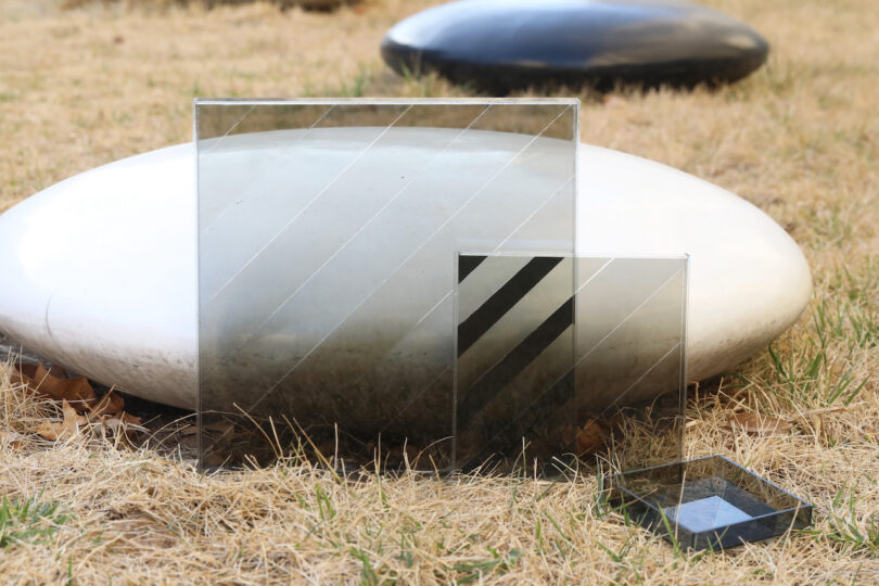 Two transparent glass panels with diagonal lines and a small glass tray are placed on dry grass in front of large, smooth, white and black stones