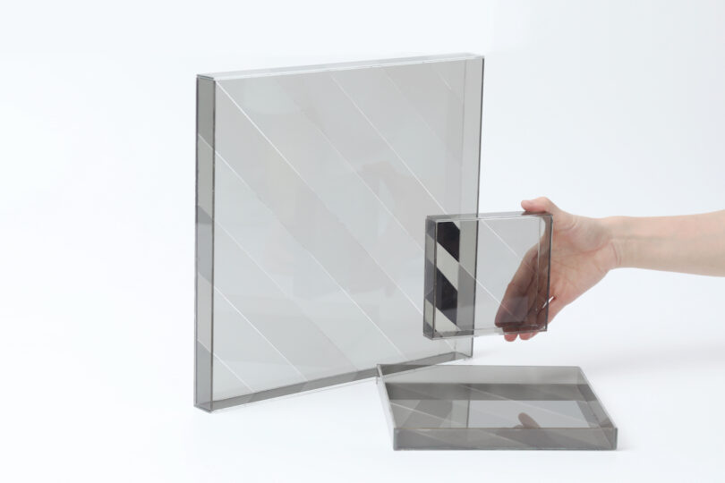 A hand holds a small transparent square with diagonal lines, next to a larger matching square and another square lying flat on a white surface