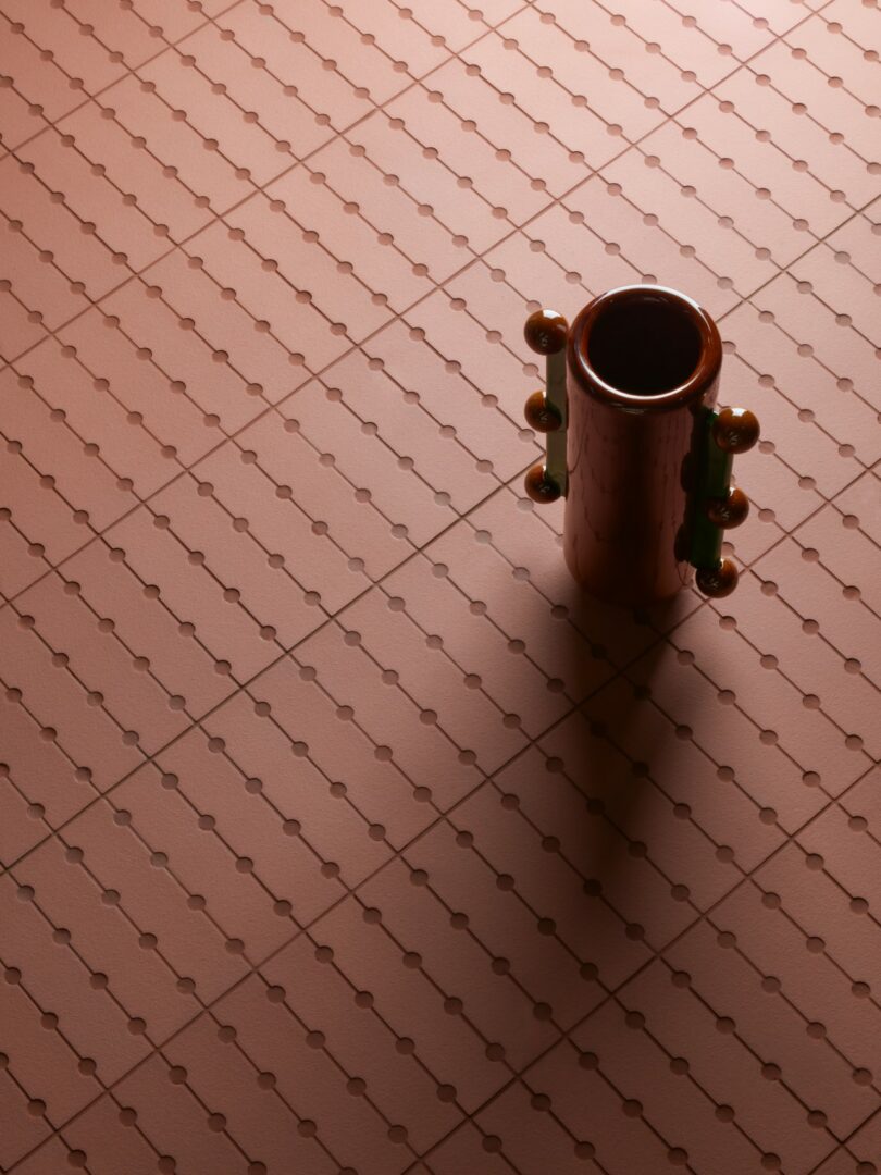 Un jarrón cilíndrico con asas redondas se alza sobre una superficie estampada de color rosa y proyecta una sombra hacia la derecha.