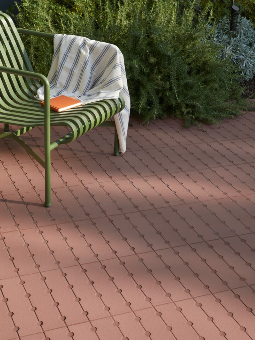 Una silla de metal verde con tela a rayas en los reposabrazos y un cuaderno naranja colocado en una terraza cubierta con azulejos rojos entrelazados, junto a plantas verdes