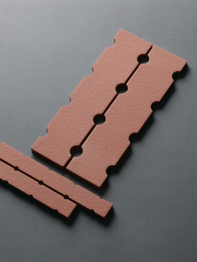 Tres azulejos rectangulares texturizados de color marrón rojizo con huecos entrelazados y recortes circulares dispuestos sobre una superficie gris