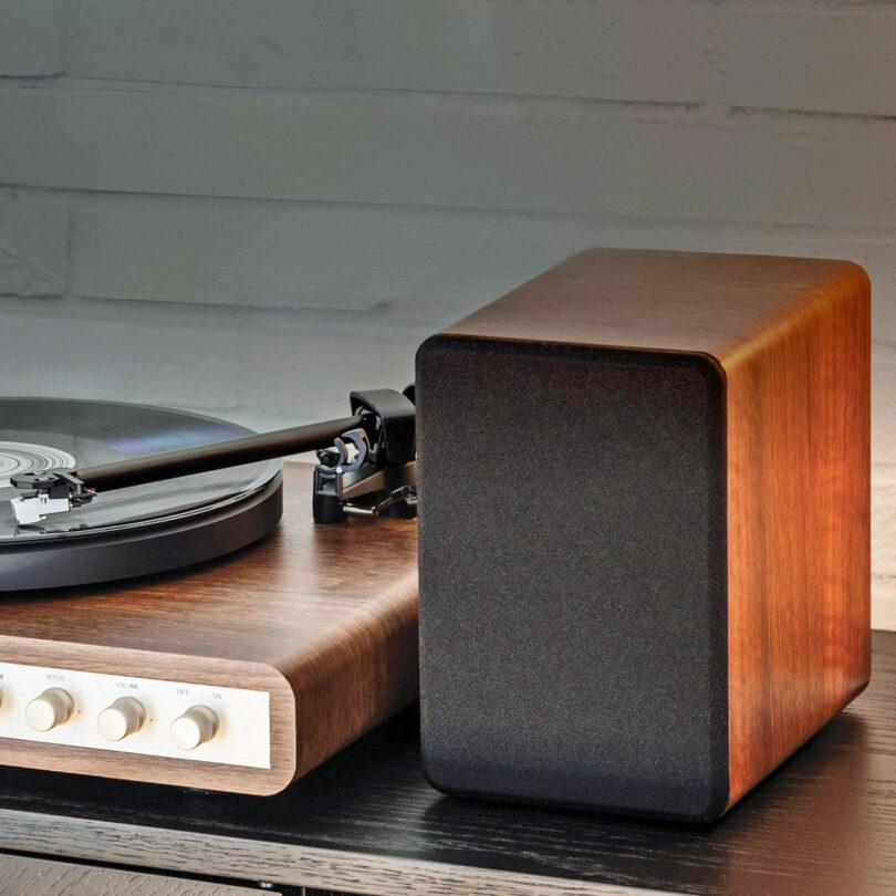 A wooden Crosley C65 turntable with a clear dust cover, playing a vinyl record, sits on a dark cabinet next to matching speakers.