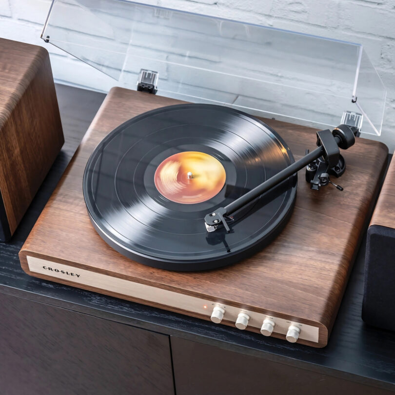 A close-up of a Crosley C65 wooden turntable playing a vinyl record next to a matching wooden speaker on a dark surface against a light brick wall.