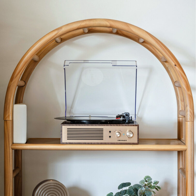 A Crosley Miles wooden record player with a clear lid sits on a beige coffee table, playing a vinyl record.