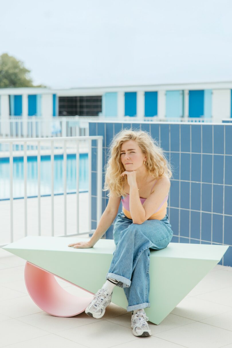 A woman with curly blonde hair sits on a geometric bench near a pool, resting her chin on her hand and looking thoughtful. She wears jeans, sneakers, and a pastel tank top