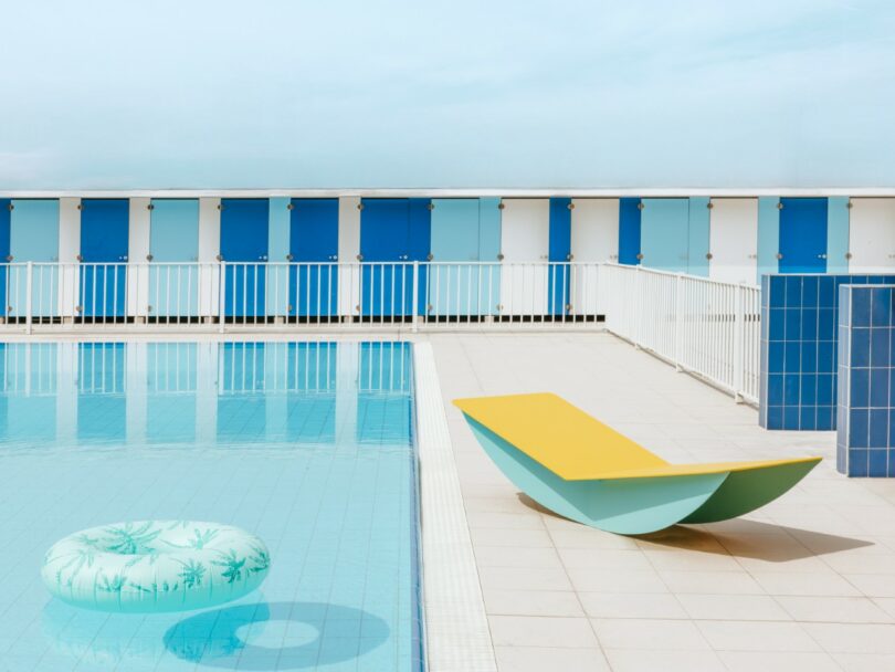A poolside scene with a blue and white inflatable float in the water, a modern yellow and blue lounge chair, and a row of blue lockers in the background