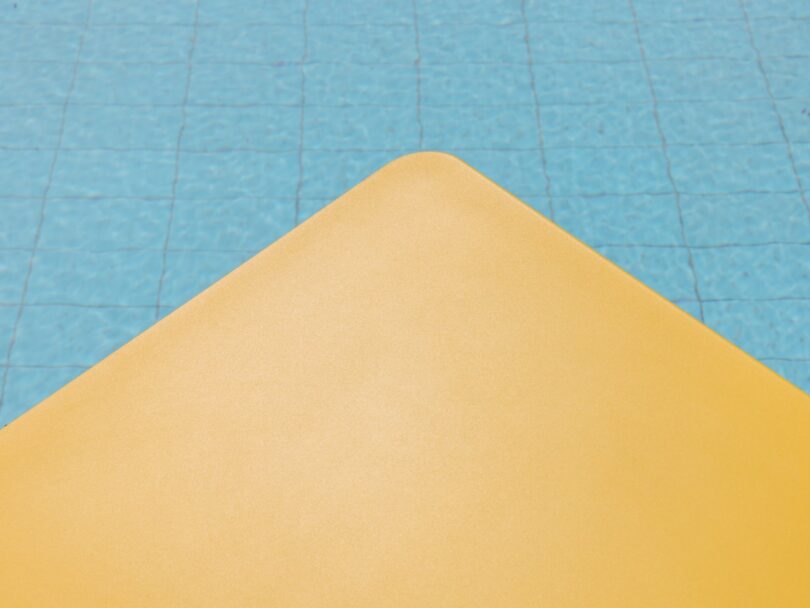 A yellow surface with a rounded corner is in the foreground, with a tiled blue swimming pool in the background
