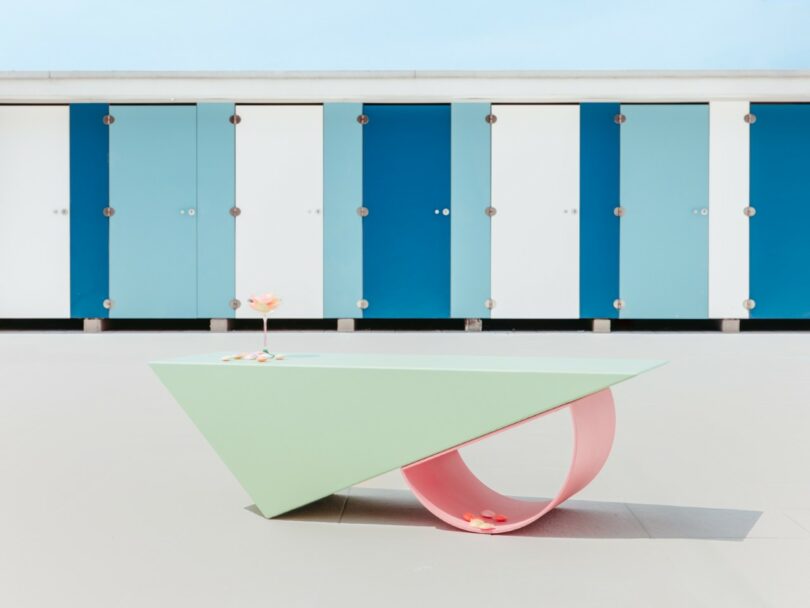 A geometric pastel green and pink table stands in front of a row of blue and white changing room doors; two cocktails and fruit slices are on the table