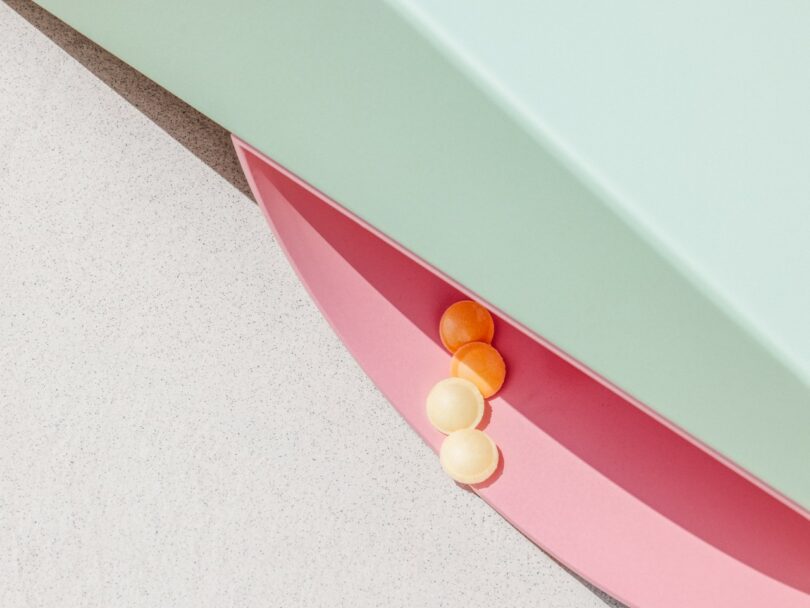 Four small spheres, two orange and two white, rest on the edge of a pink semicircular surface beneath a mint green and off-white structure under bright light