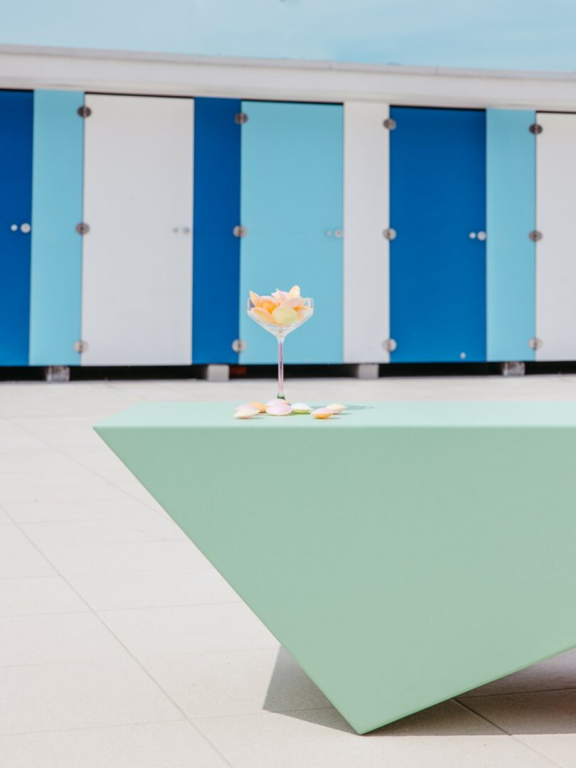 A pastel green geometric table with a cocktail glass of fruit and petals, set outdoors in front of blue and white changing room doors